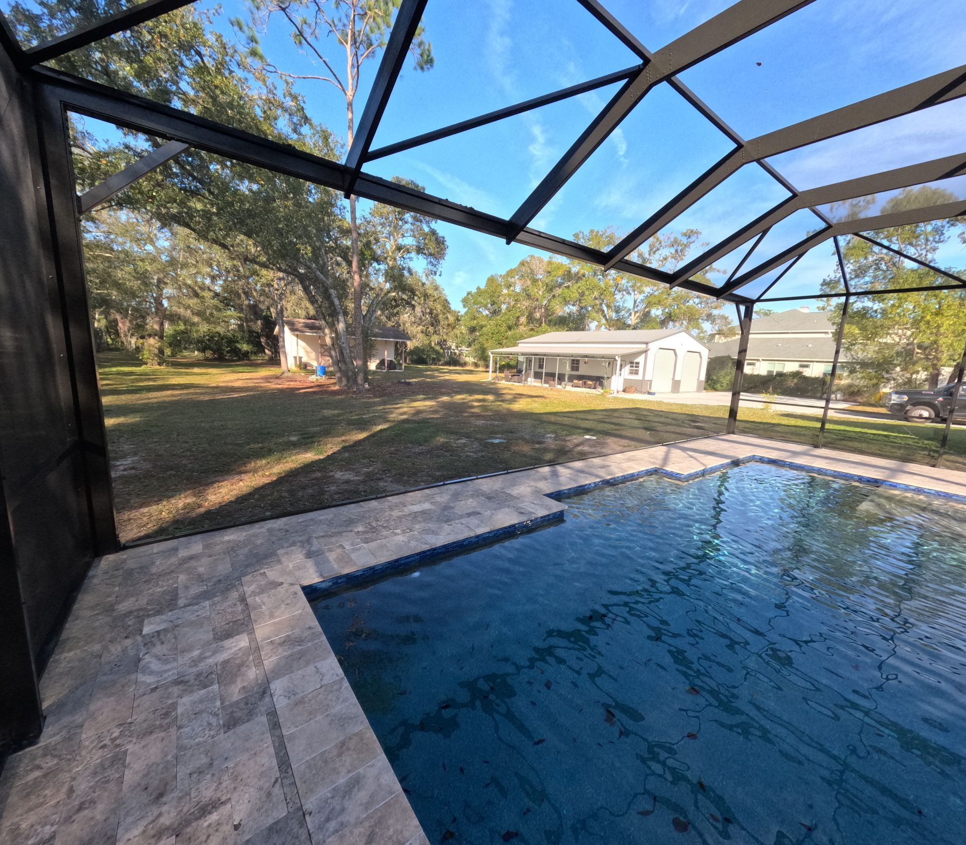 Pool with clear water, screen enclosure, and view of a grassy yard with trees and a house on a sunny day.