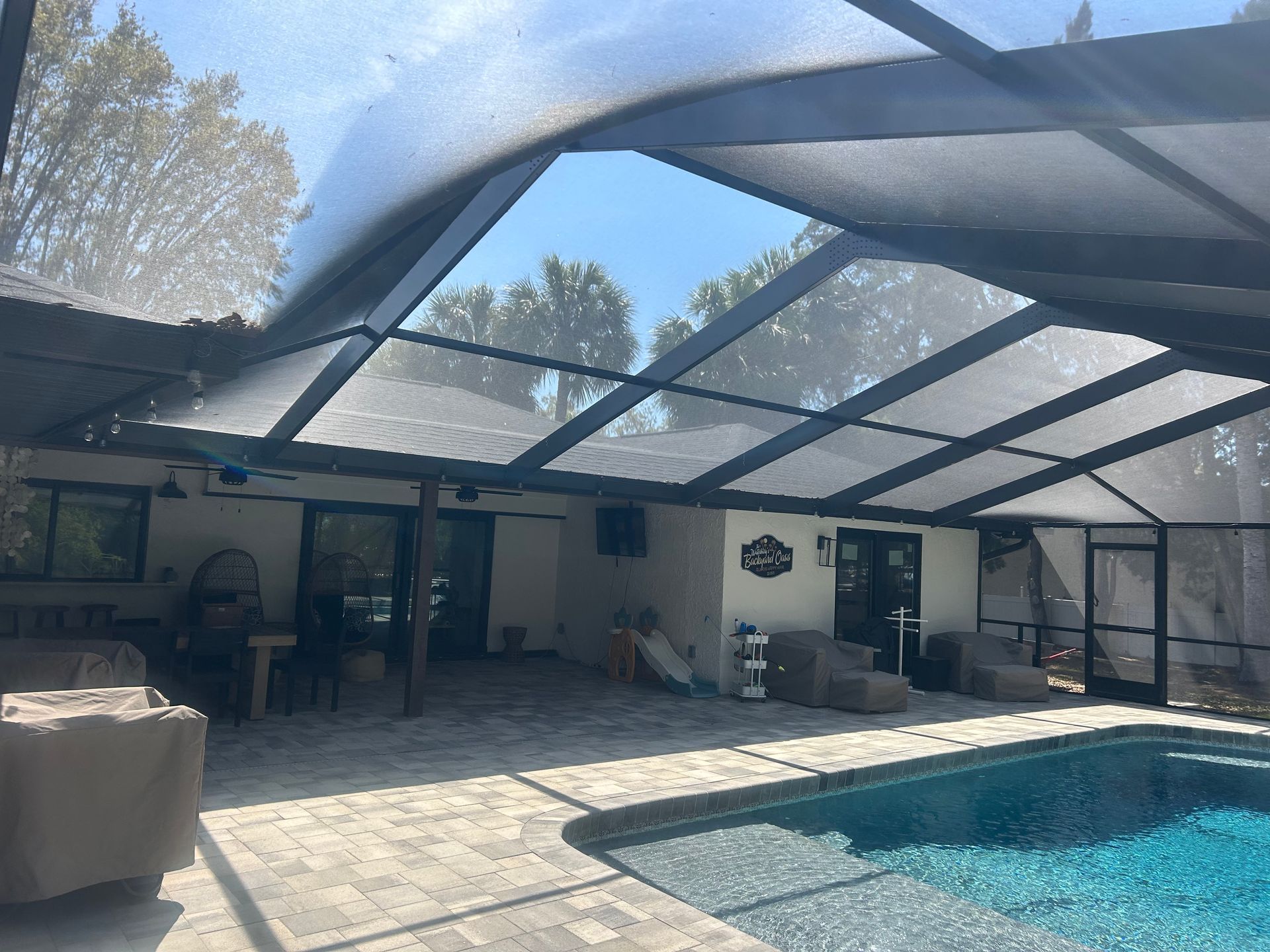 Pool patio with screen enclosure, sunny day. Blue pool, black frame, gray pavers, and a white building.