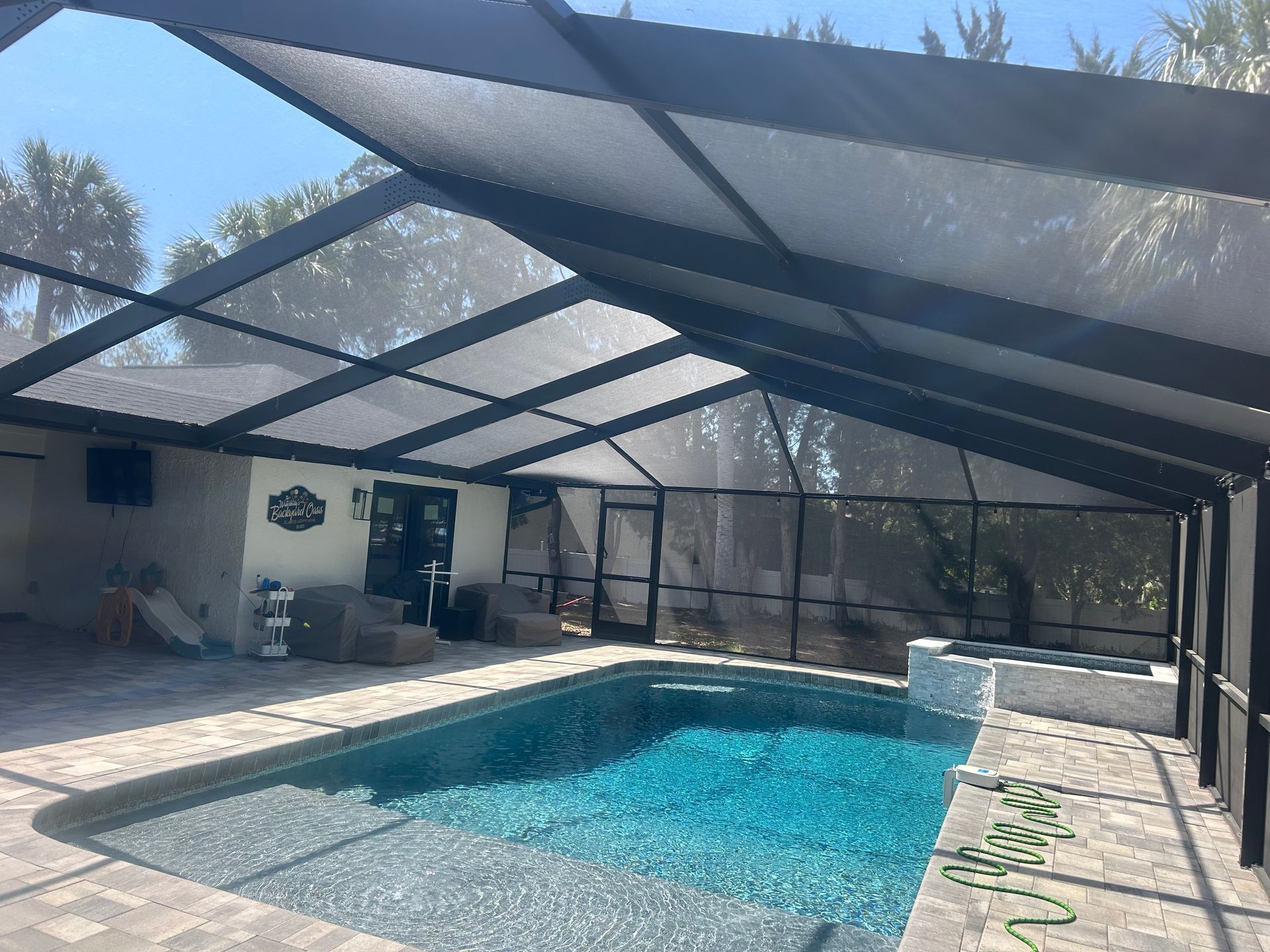 Pool enclosed by a black screen structure with a spa, on a patio, under a blue sky.