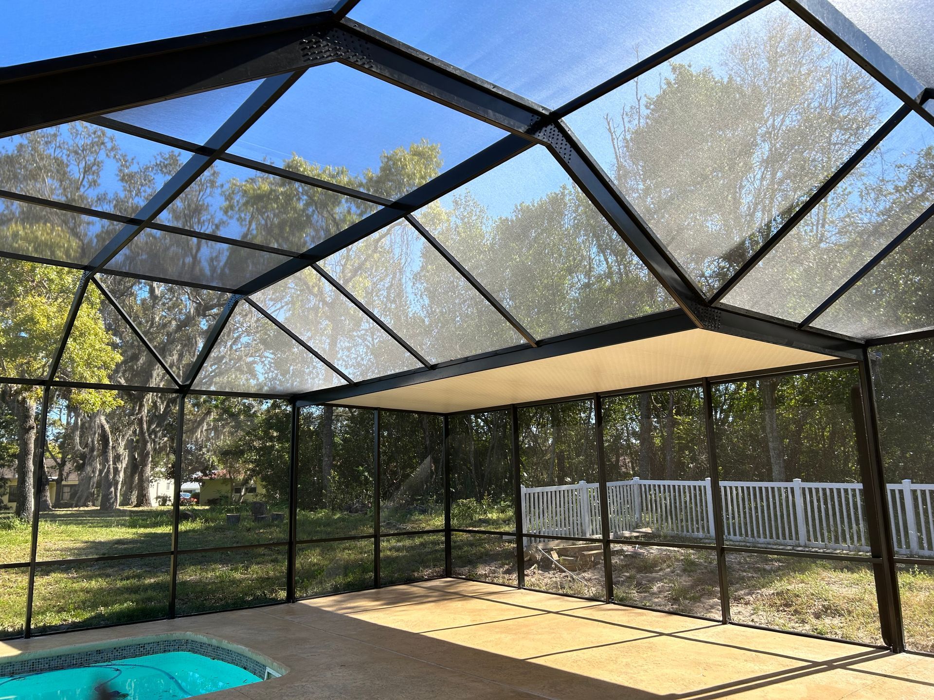 Screened-in patio with pool, black frame, clear screens, and tan ceiling panel on a sunny day.