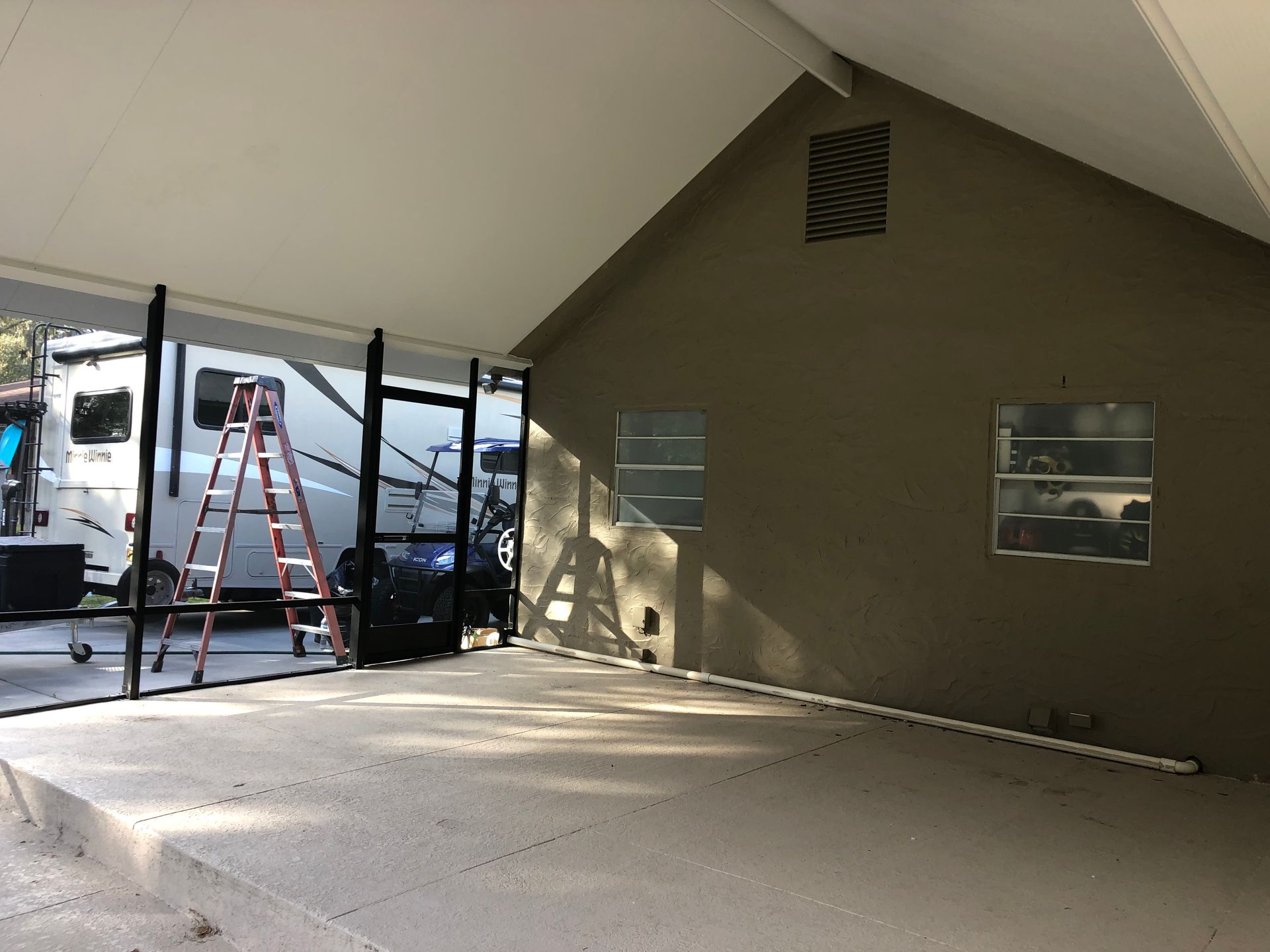 Covered outdoor area with RV visible. A ladder leans against black framed screening, and there is a cement floor.
