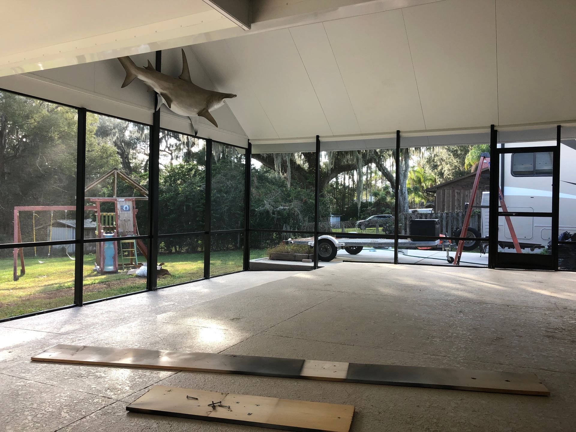 Interior view of a screened porch, with a shark mounted on the ceiling. Concrete floor, windows, and surrounding trees.
