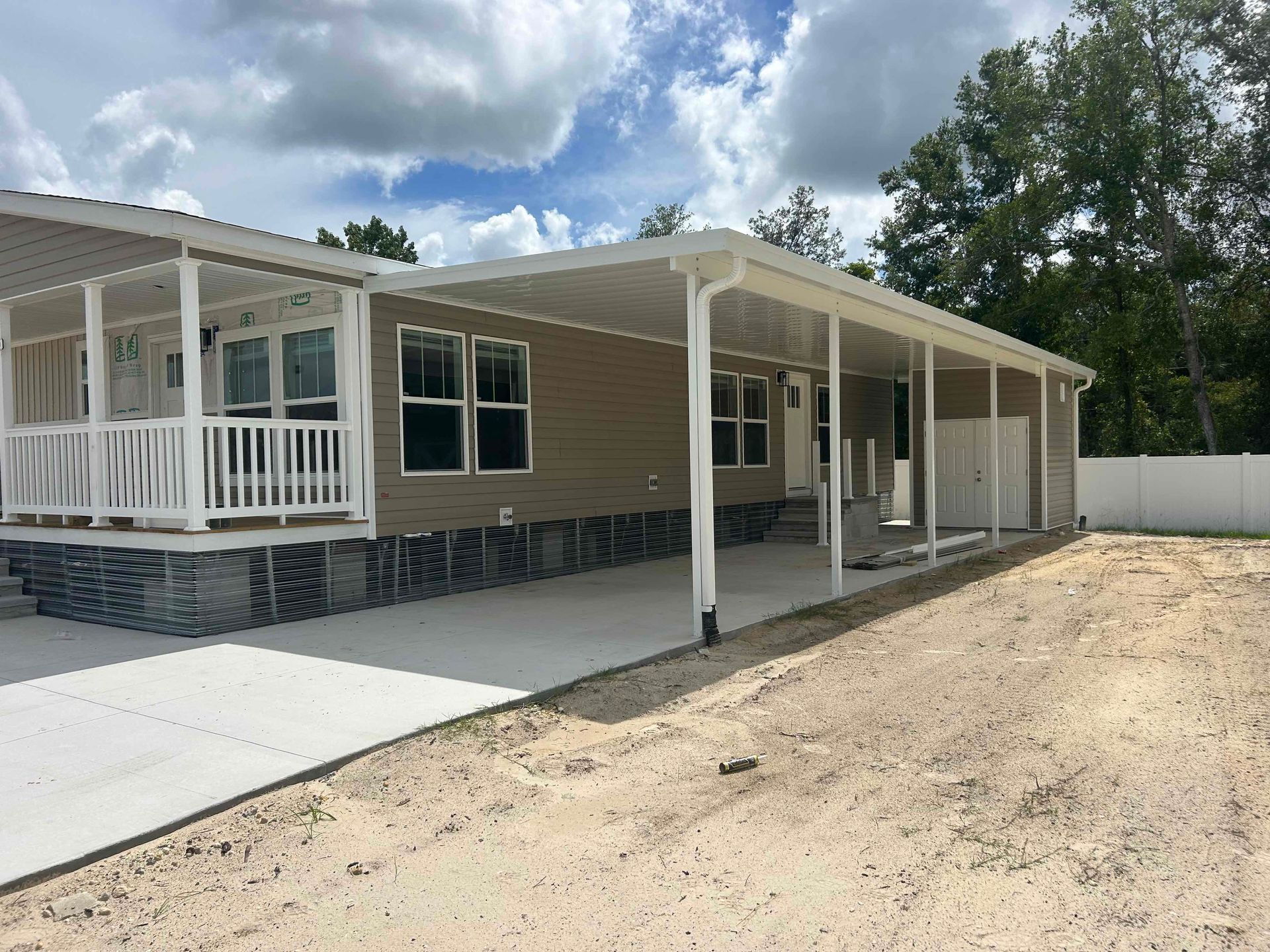 Tan house with white porch and carport, concrete driveway, set against a blue sky with clouds.