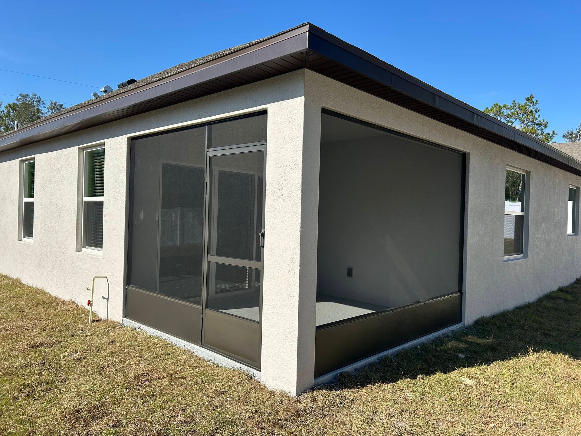Screened-in porch on a stucco house with dark brown trim and screen. Sunny outdoor setting with grass.