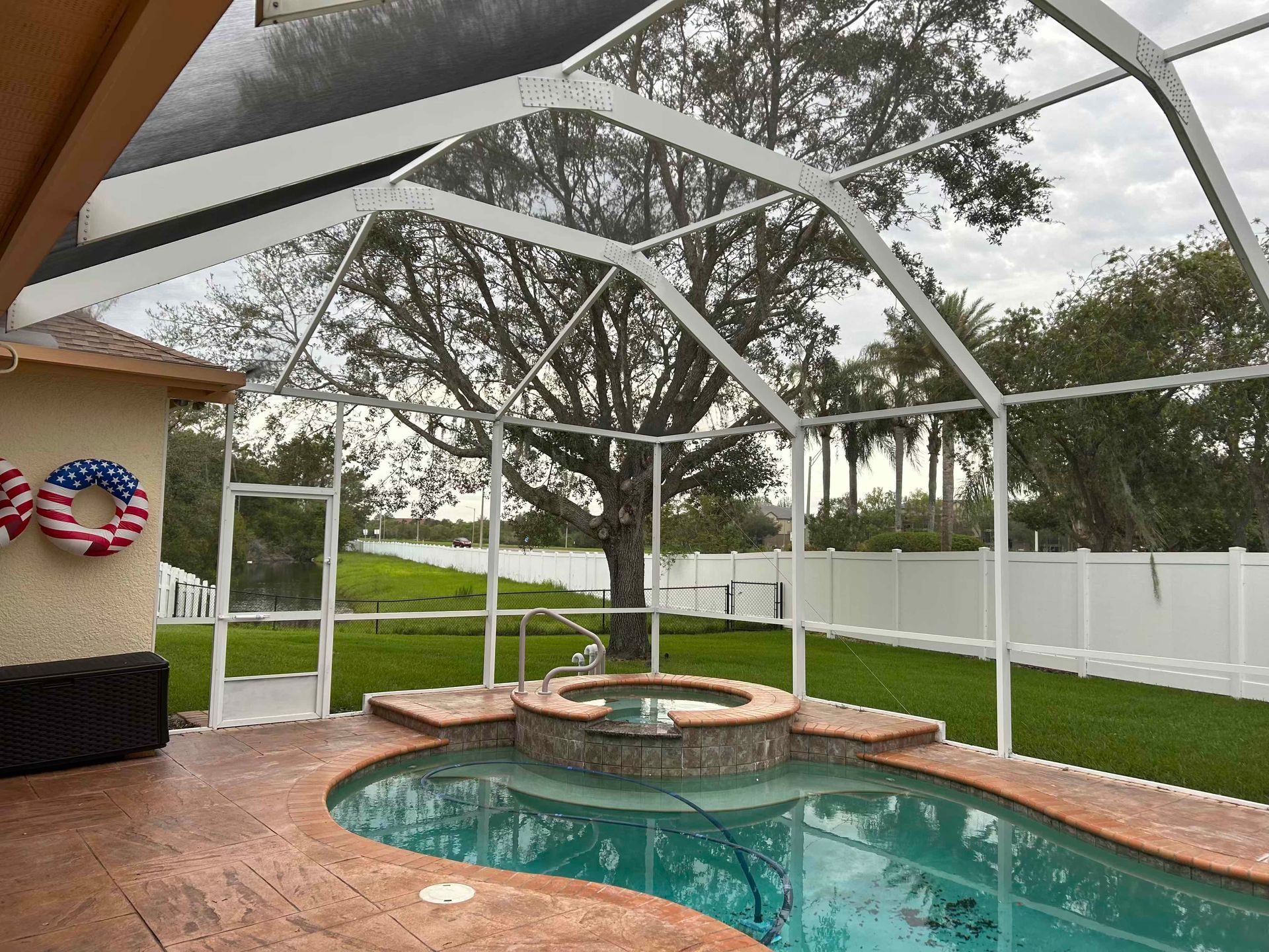 Pool enclosure with pool, spa, and backyard view. White framing, brown pool deck, green grass, overcast sky.