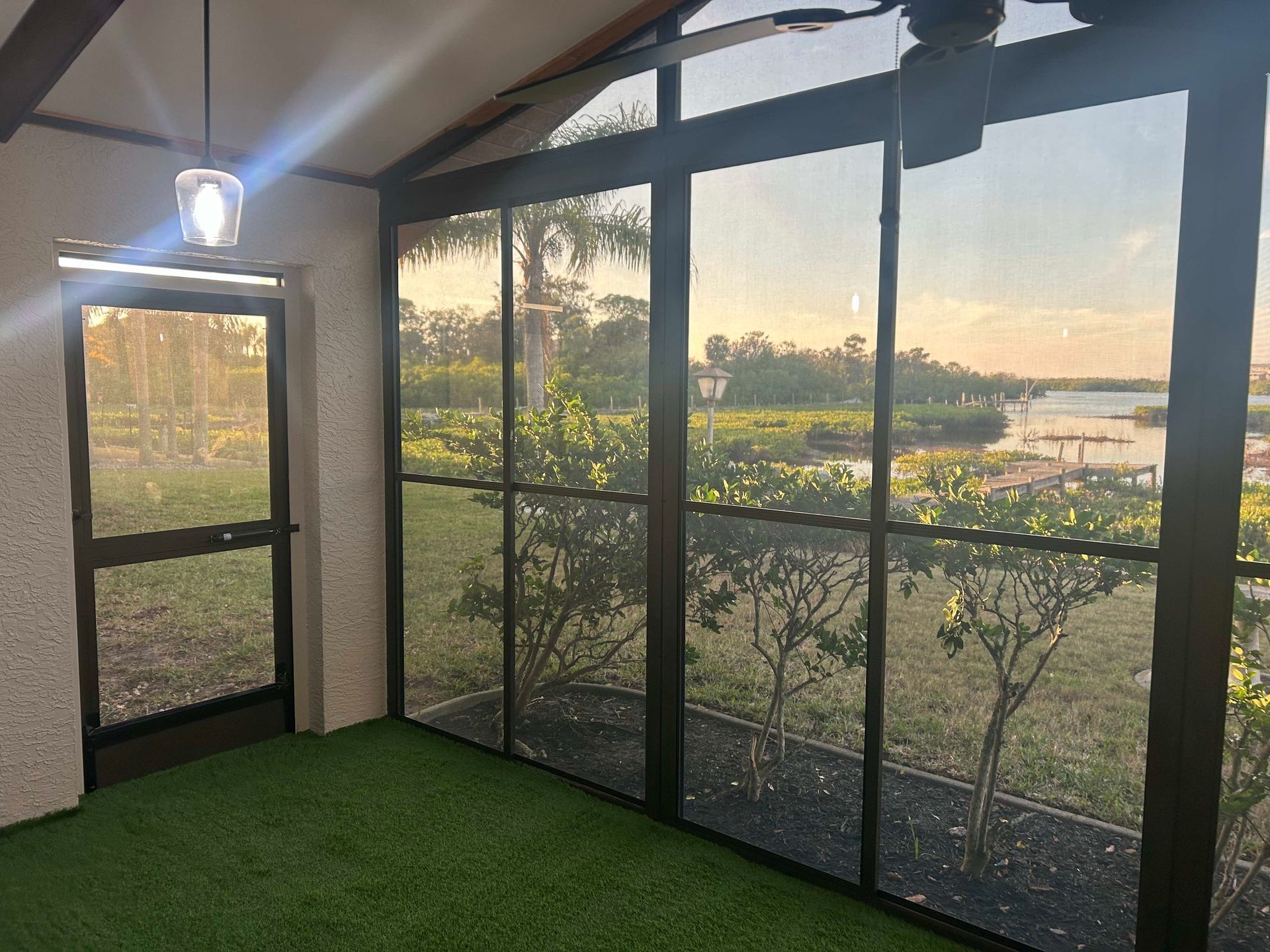 Screened-in porch with view of water and greenery. Light shines in from a door and a ceiling light.
