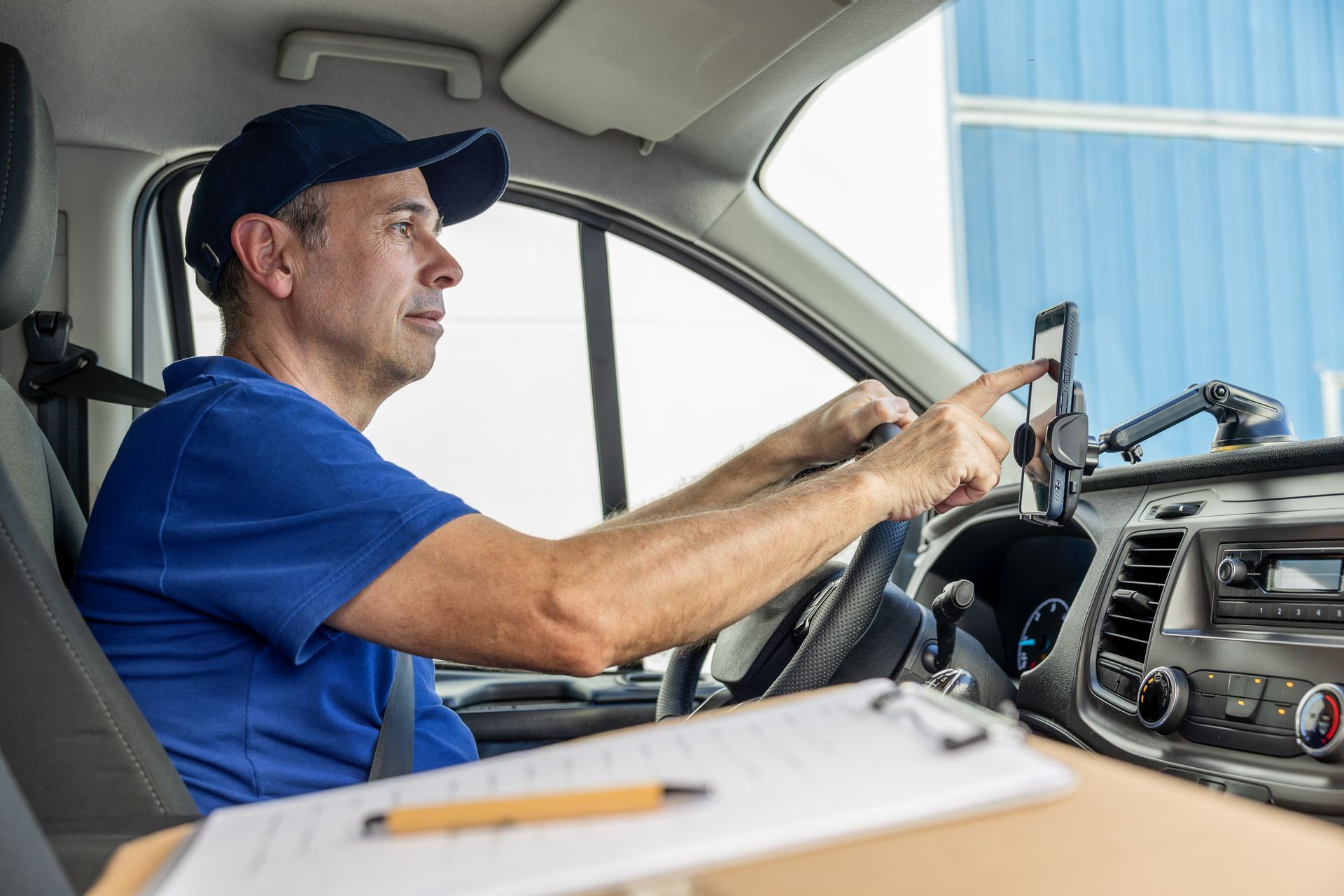 Driver in blue shirt and cap using a phone mount inside a van, with a dashboard and notebook in view