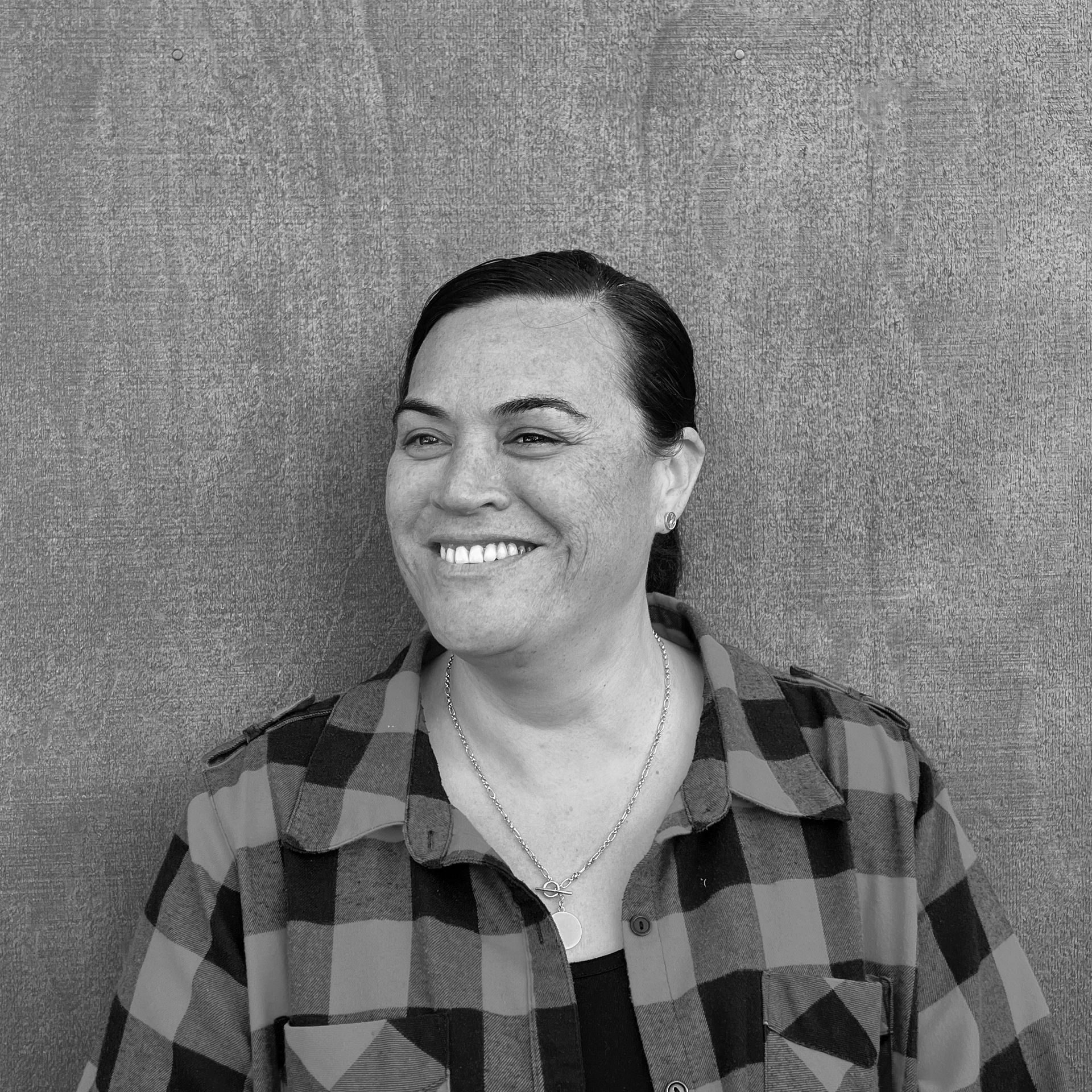 Smiling person in a black-and-gray plaid shirt standing against a textured gray wall
