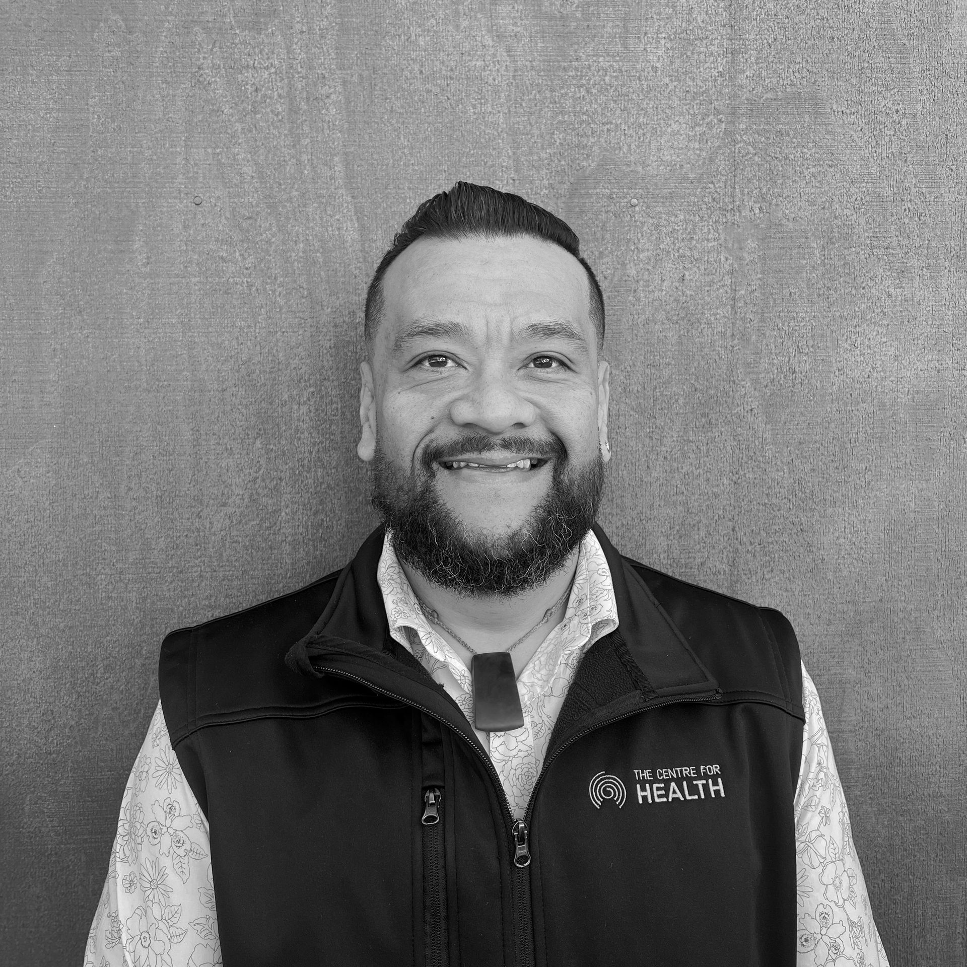 Smiling man in a dark vest standing against a plain wall, facing the camera.