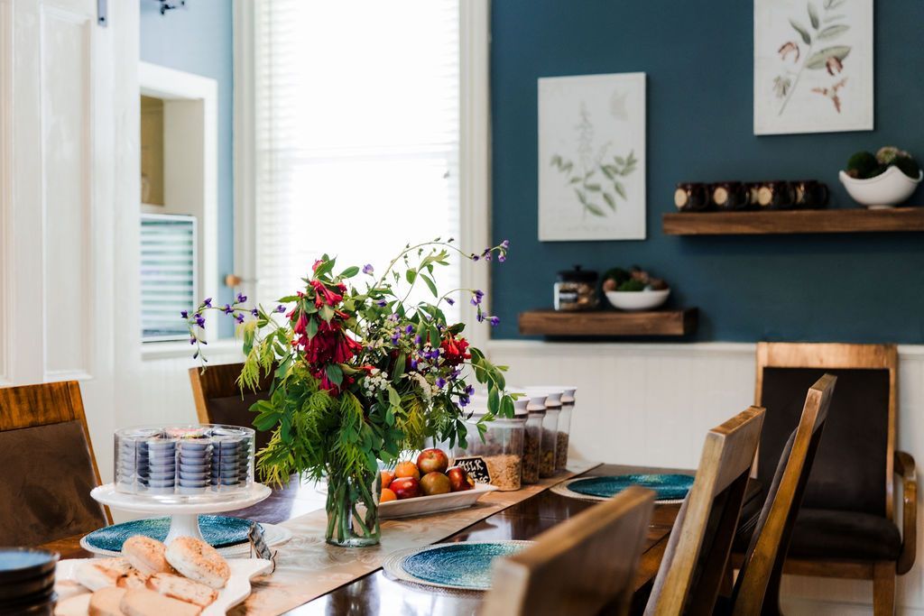 Dining room with table set for a meal, flowers as centerpiece, blue wall, wooden chairs.