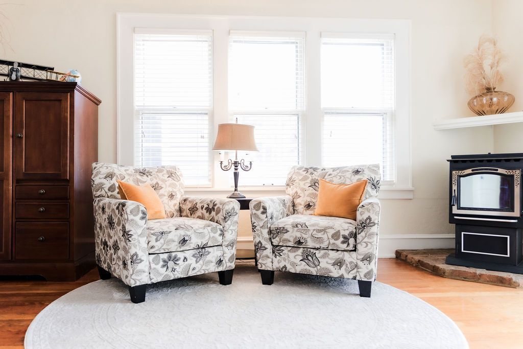 Two floral armchairs with orange pillows, a lamp, and fireplace in a bright living room.