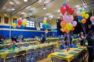 A high school gymnasium set up for an event with yellow-covered tables, chairs, and colorful balloon centerpieces.