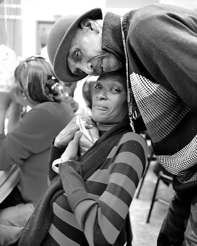 A person in a hat leans in close to another person, who is sitting and looking toward the camera in a light-filled space.