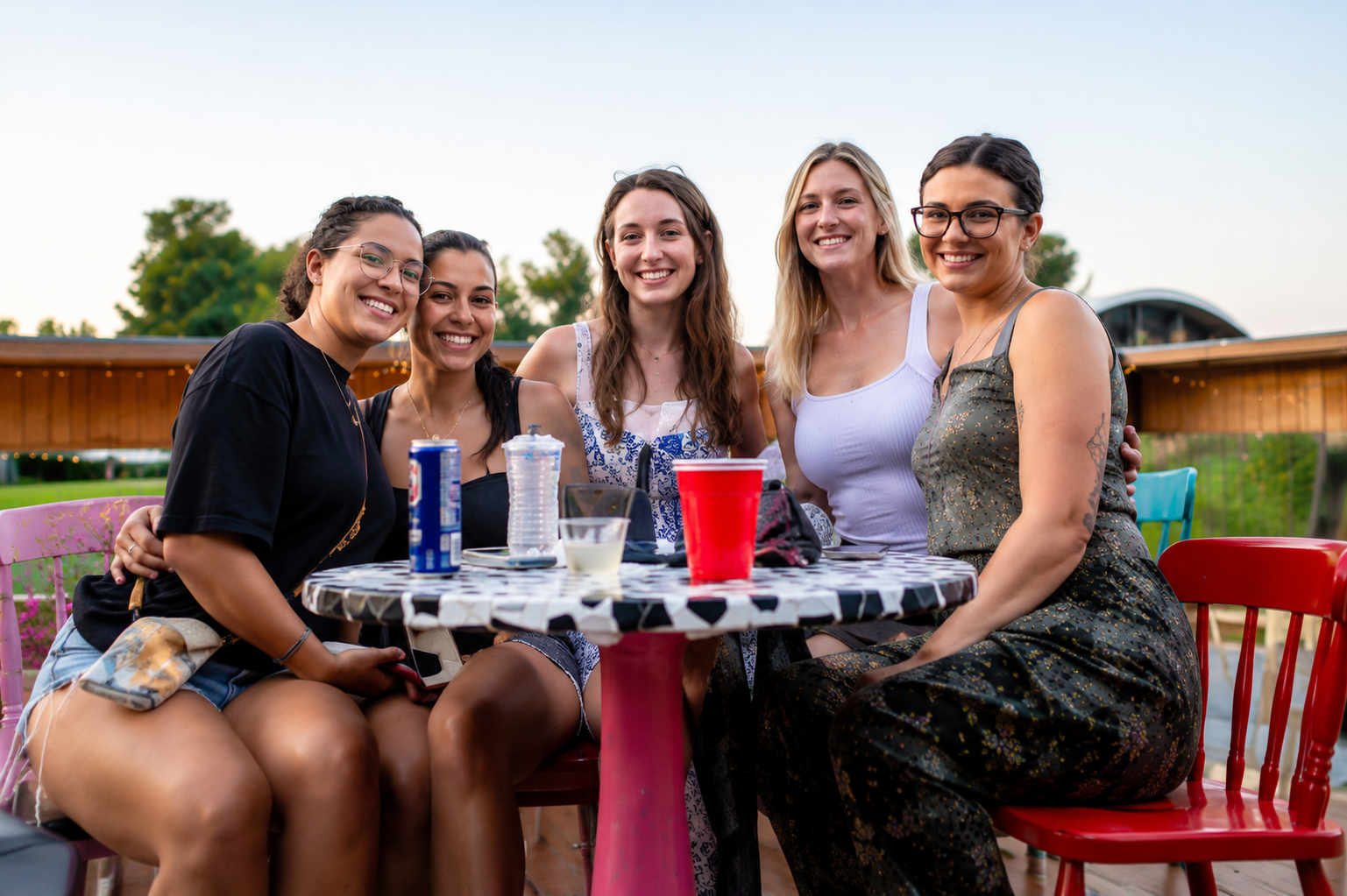 Five friends smiling at an outdoor table with drinks, seated in a grassy field at dusk