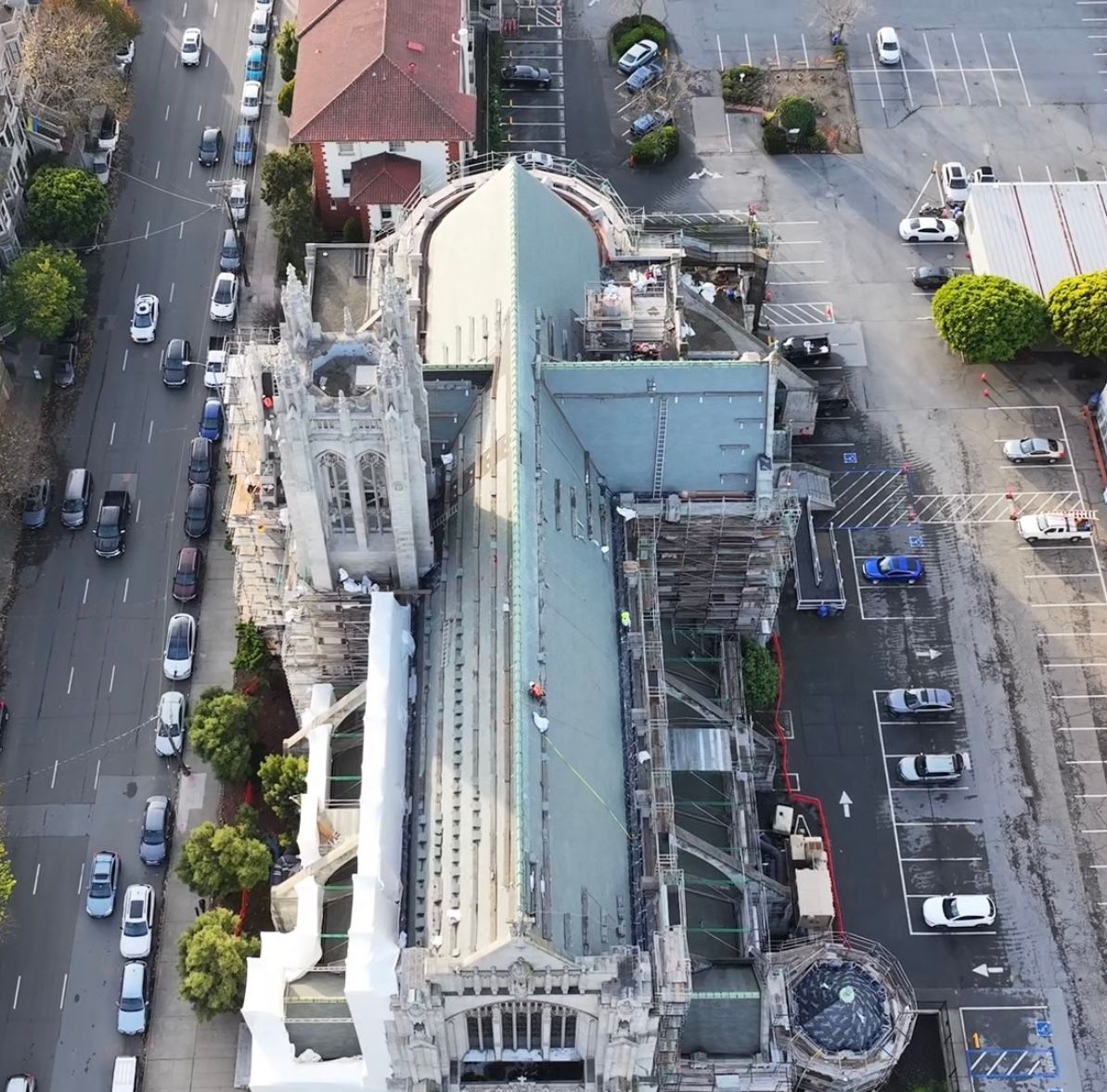 Aerial view of a cathedral with a tall tower, blue-green roof, and street with parked cars.