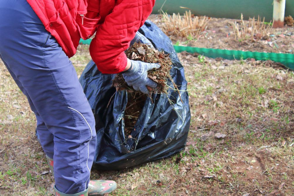 Person in Red Jacket and Blue Pants Putting Leaves Into a Black Trash Bag — All Suburbs Garden Bags in Kuranda, QLD