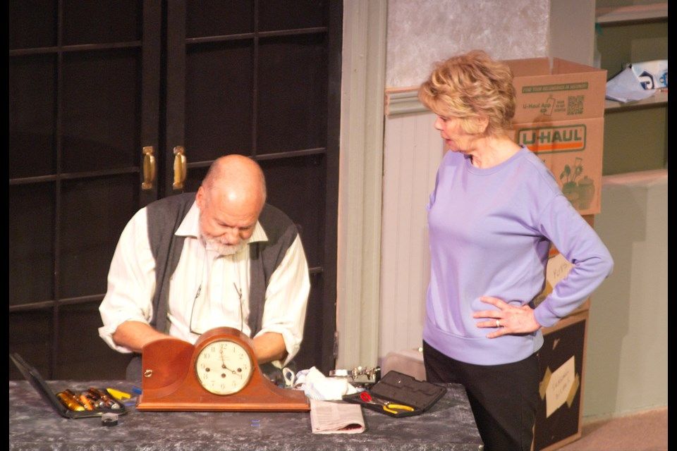 Man fixing a clock, woman watches with hand on hip; indoor setting with moving boxes.