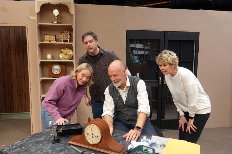 Four people examining an antique clock on a table. Setting appears to be a stage set with shelving and doors.