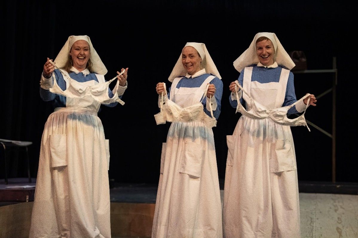 Three women in nursing uniforms hold up bras on a stage.