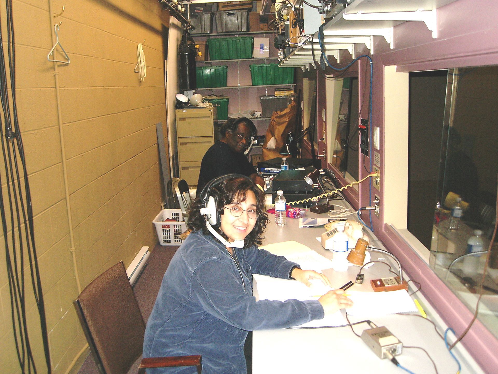 Woman wearing a headset smiles in a booth. Another person works behind her. They are in a small, windowed room.