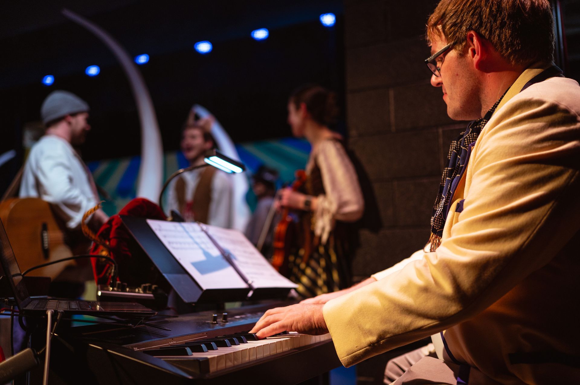 Keyboardist performing onstage; bandmates in background.