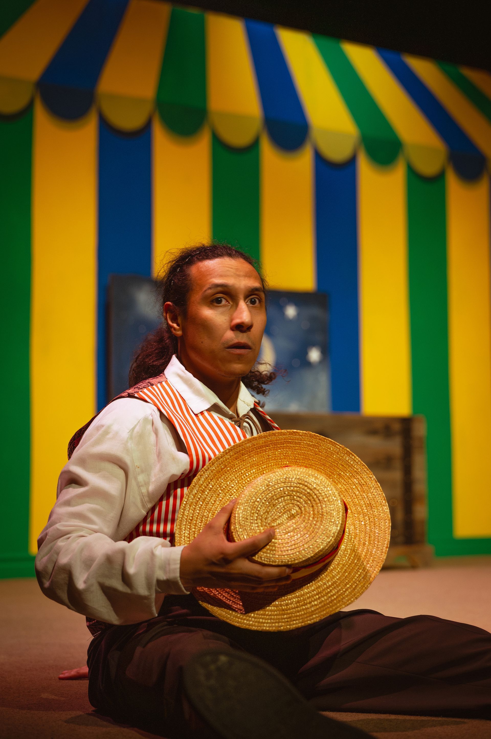 Man in vest, holding a straw hat, looks up in concern; colorful circus tent backdrop.