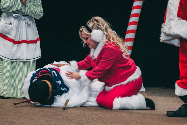 Woman in pink Santa suit kneels next to a snowman on the floor. Stage backdrop.