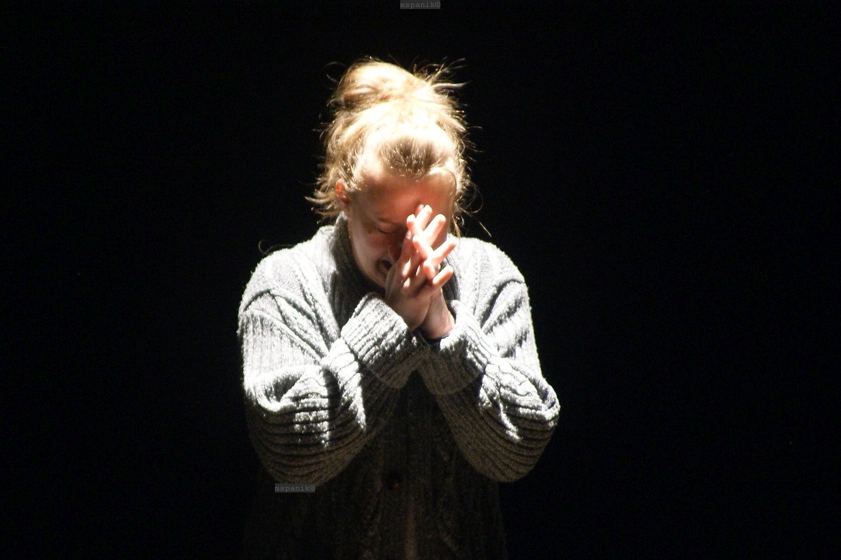 Woman in shadow, hands clasped, head bowed. Dark background, single light source.