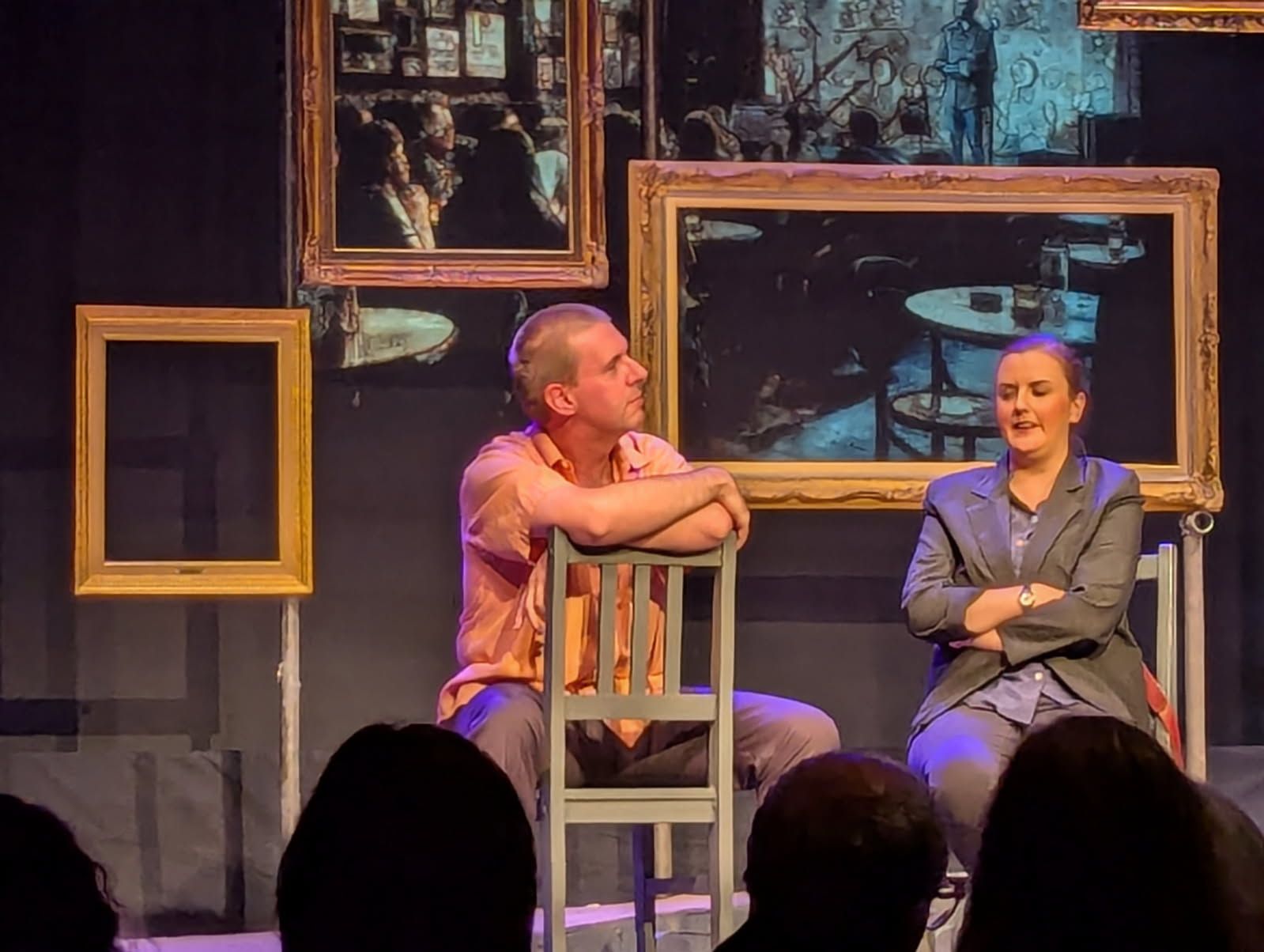 Two people on stage, framed by empty and full paintings. One sits, arms on a chair, the other sits with arms crossed.