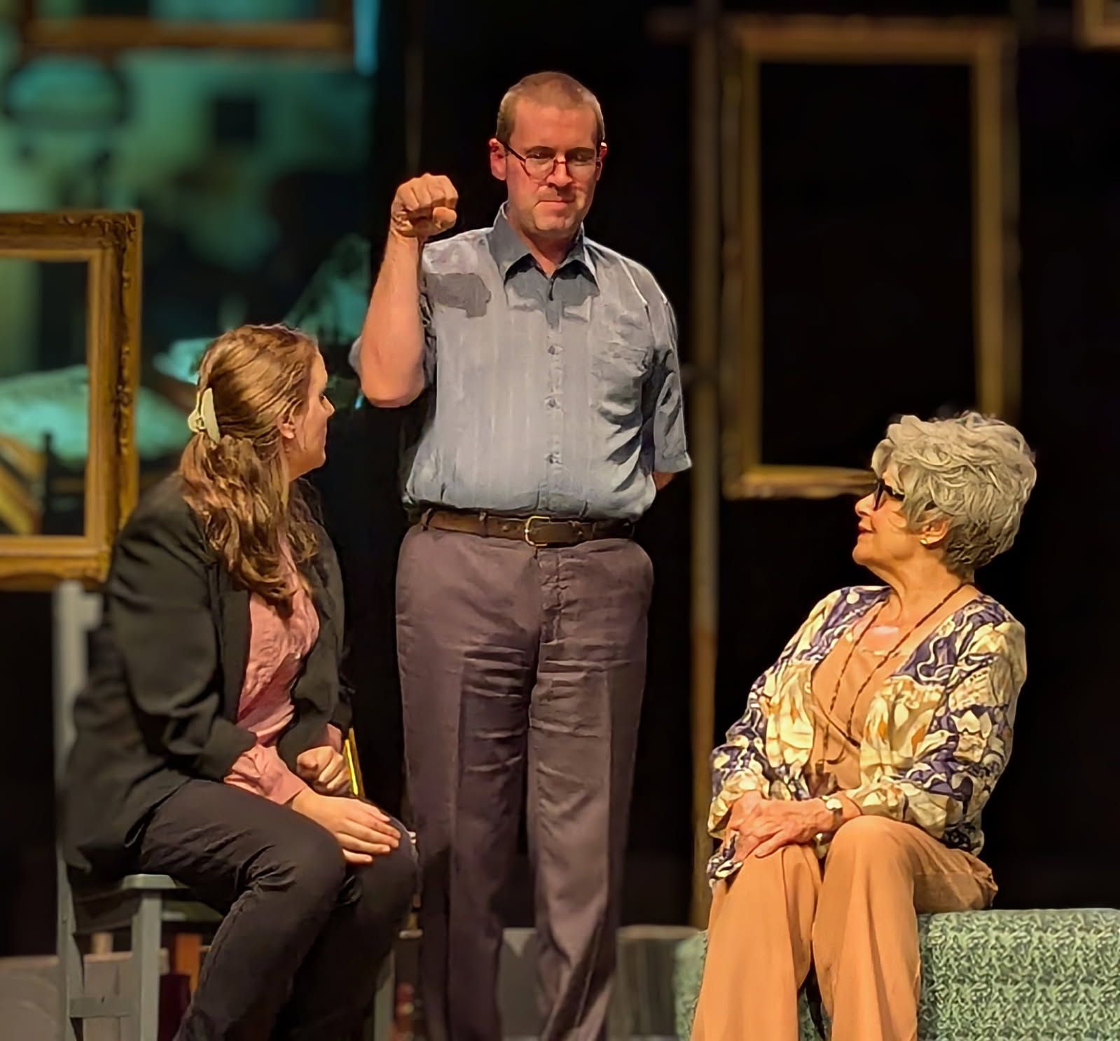 Three people on stage. Man raises fist. Woman in blazer sits. Woman in floral top sits. Frames in background.