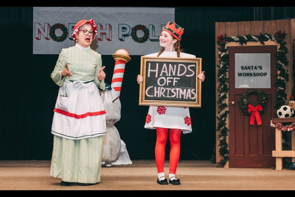 Two people in costume on a stage: one in a dress and apron, the other holding a sign that says 