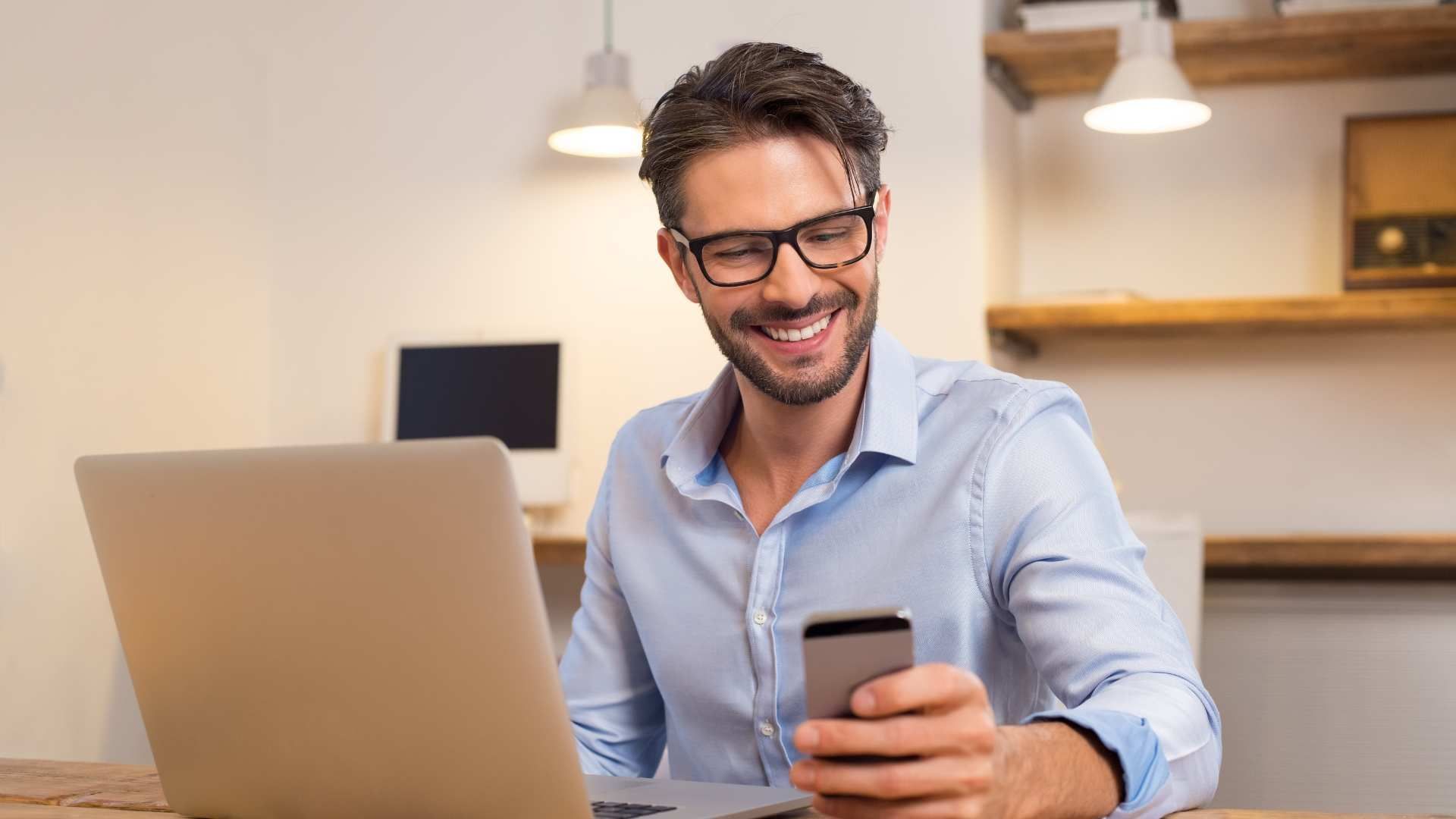 Smiling man in glasses using a laptop and smartphone at a desk in a bright home office