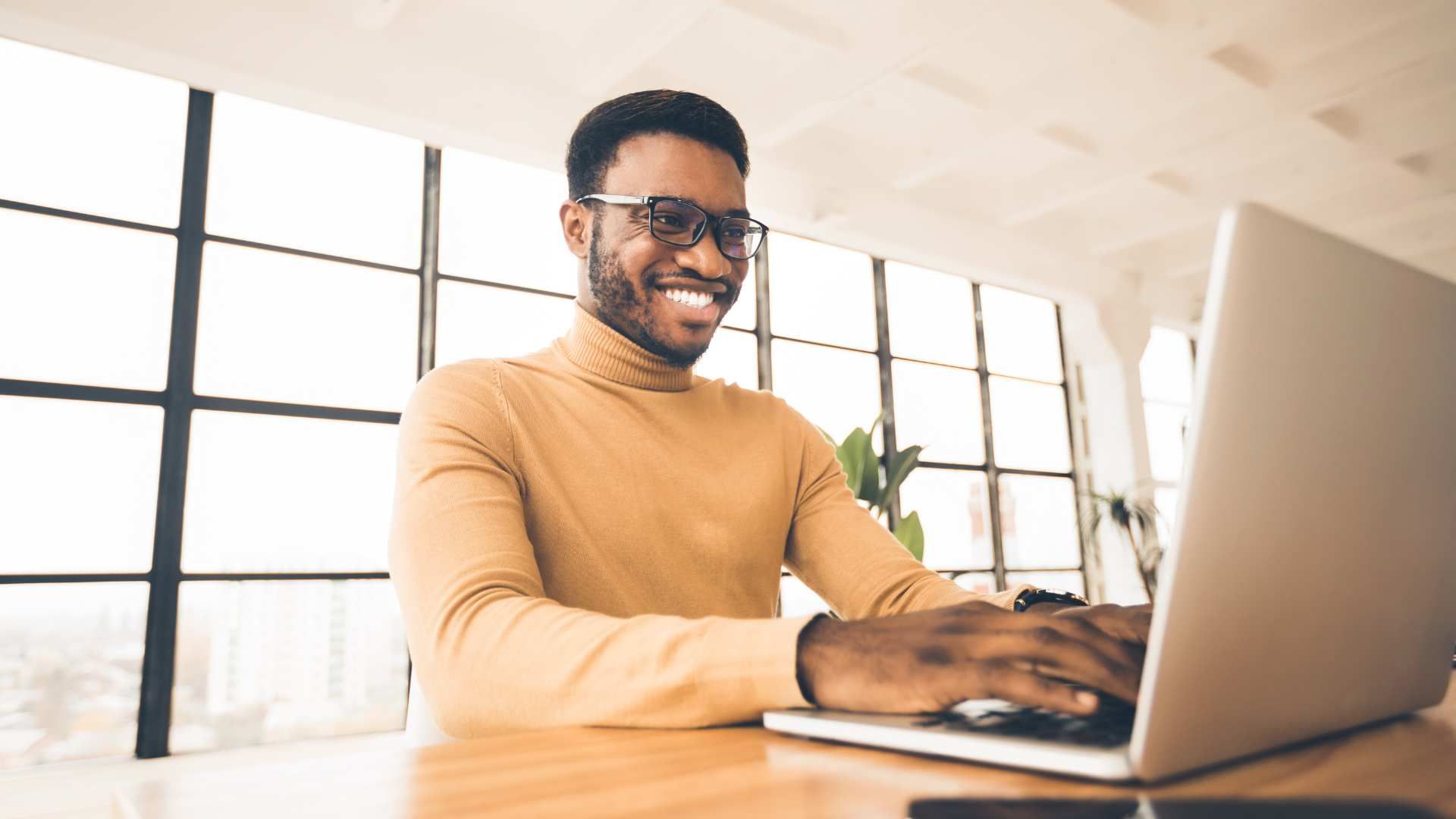 Smiling person in a tan sweater typing on a laptop in a bright office.