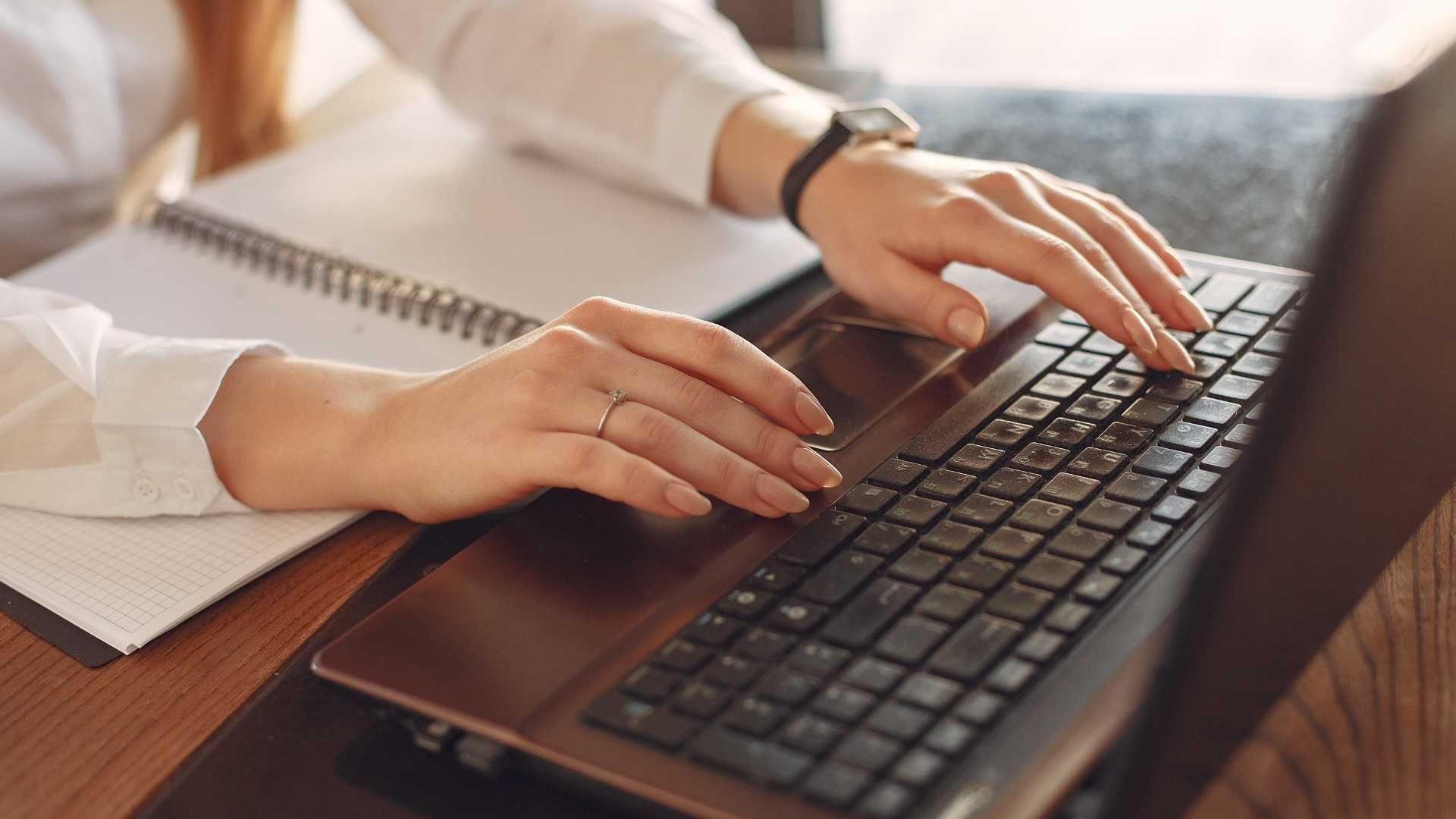 Hands typing on a laptop keyboard beside an open notebook on a desk