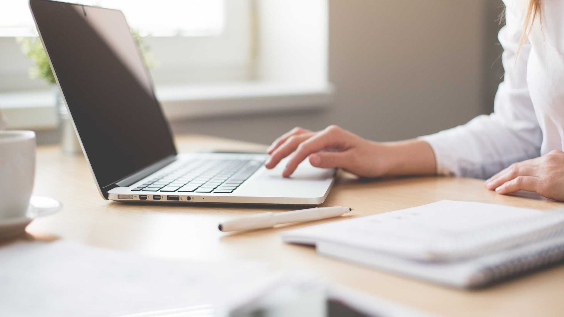 Person typing on a laptop at a bright desk beside an open notebook