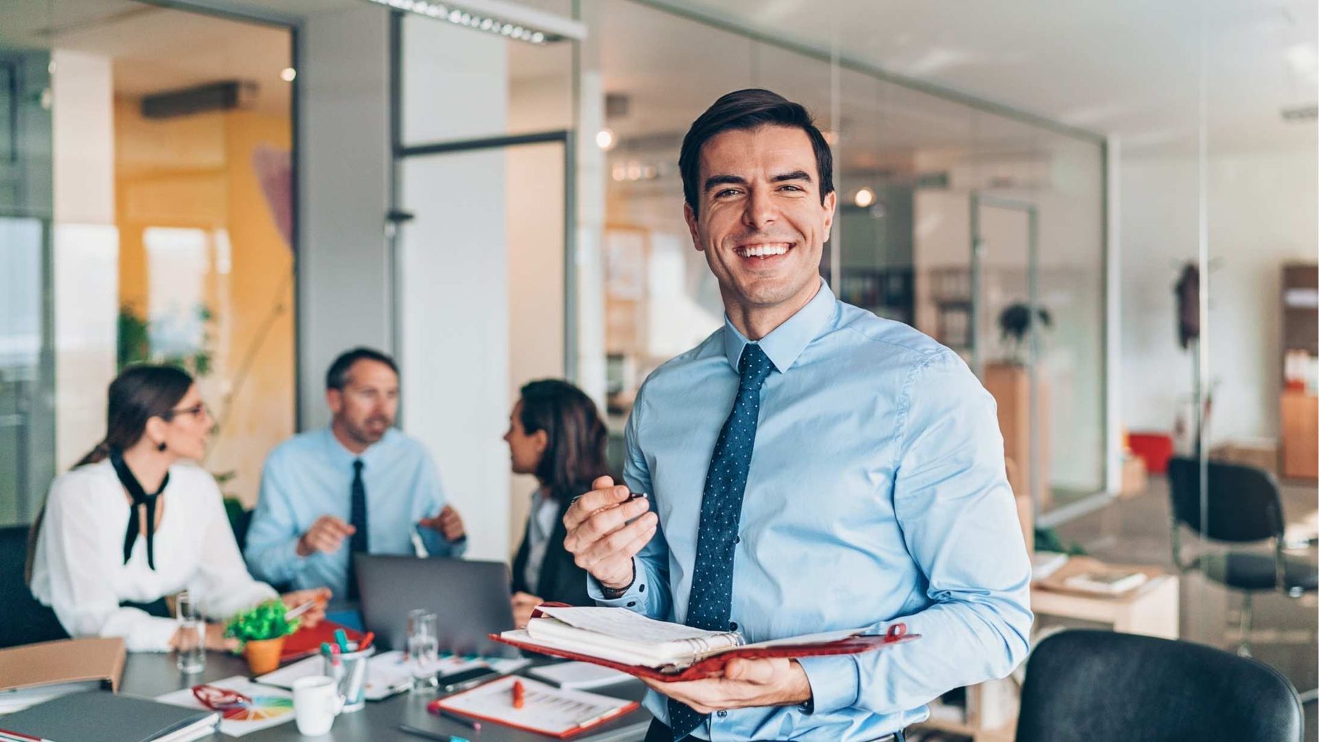 Smiling man in a blue shirt and tie holding a tablet in a glass-walled office meeting room
