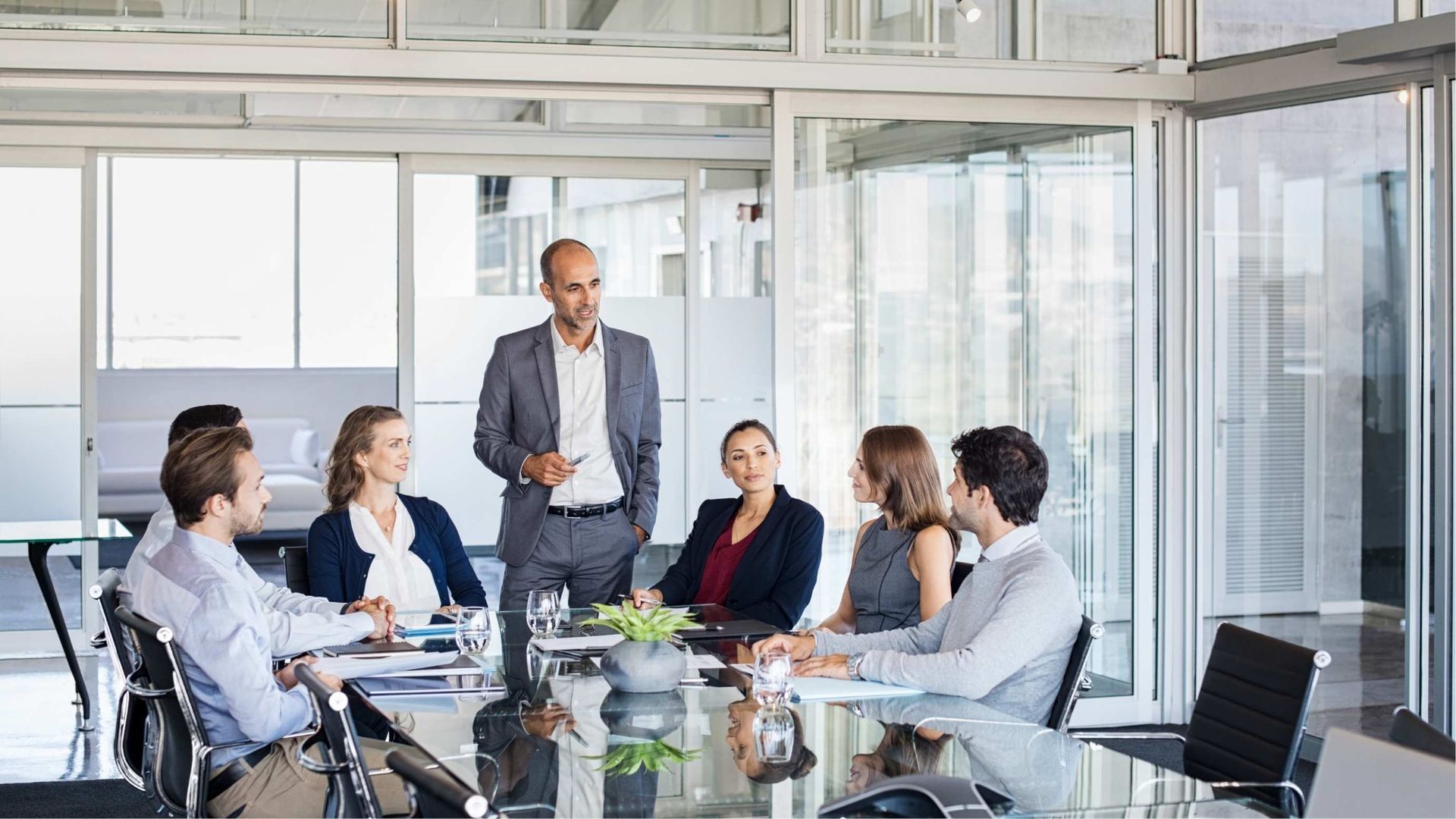 Business meeting in a glass conference room with a presenter standing beside seated colleagues around a table