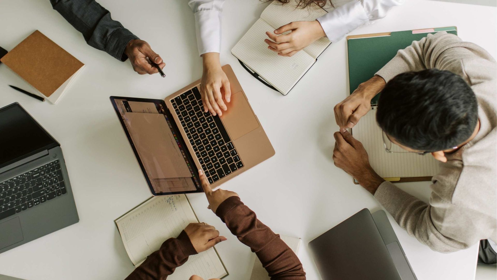 Overhead view of people gathered around a laptop, collaborating at a table with notebooks and coffee cups