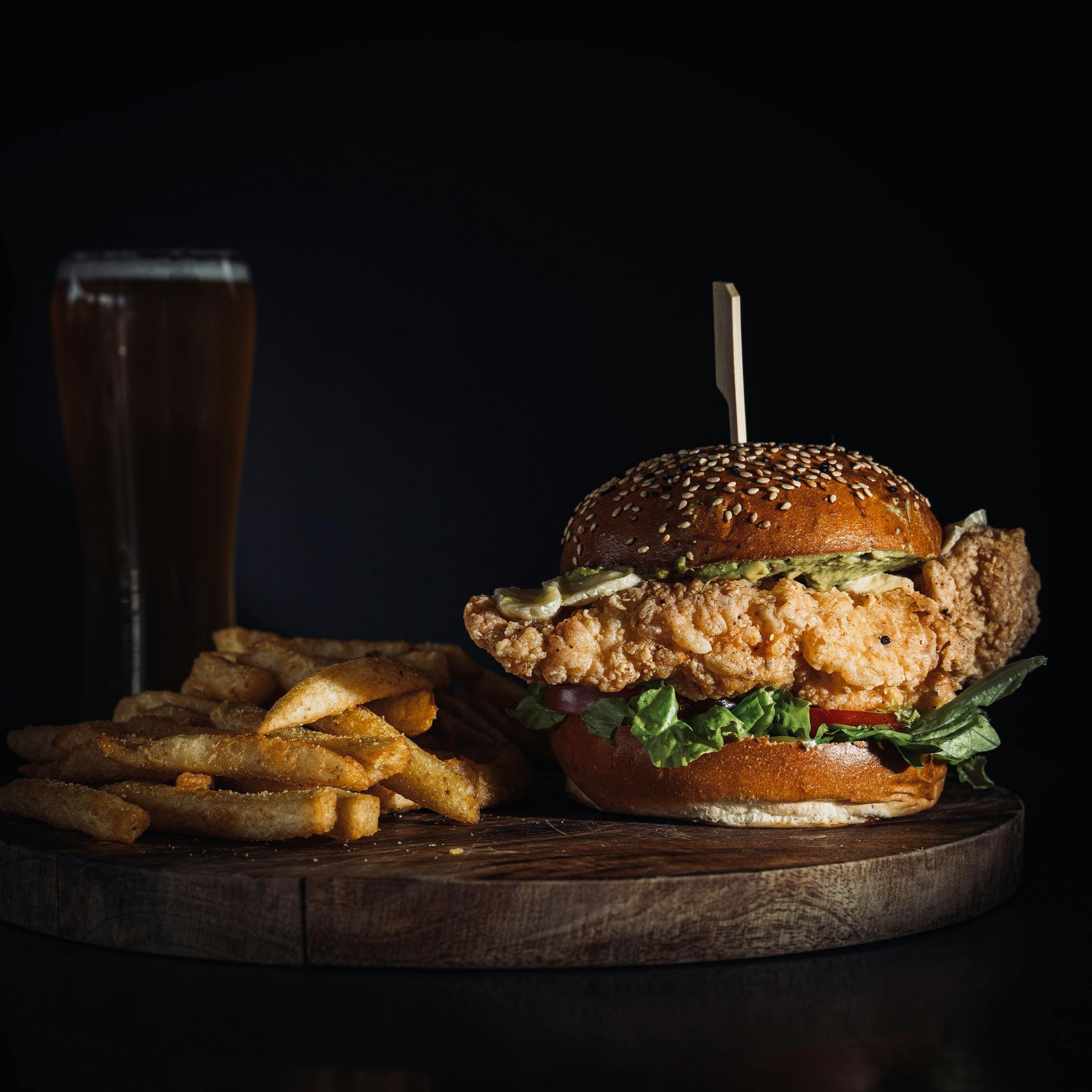 A chicken sandwich and french fries on a wooden cutting board next to a glass of beer.
