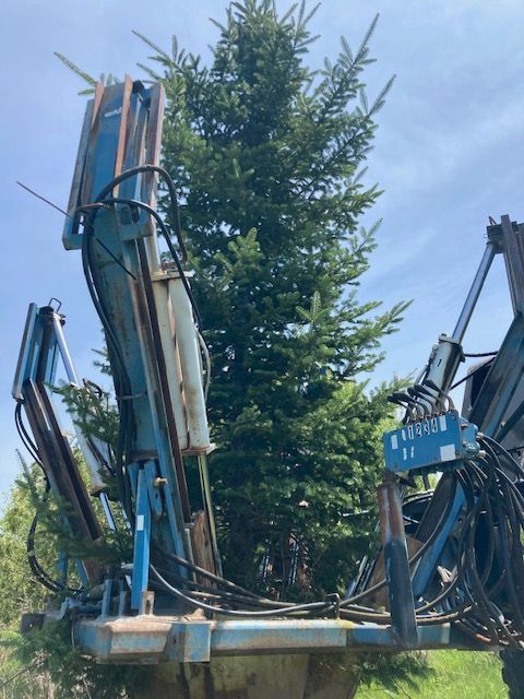 A blue tree spade machine transporting a large evergreen tree against a blue sky.