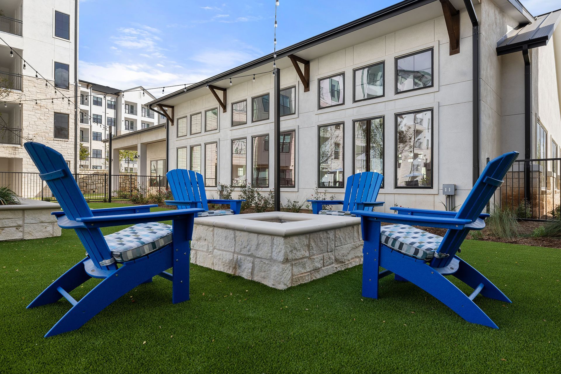 Blue Adirondack chairs surround a stone fire pit on green turf, next to a white building with windows at The Wyatt, offering Cedar Park luxury apartments.