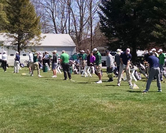 A group of people wearing casual clothing and hats practice swinging golf clubs on a sunny outdoor grass driving range.
