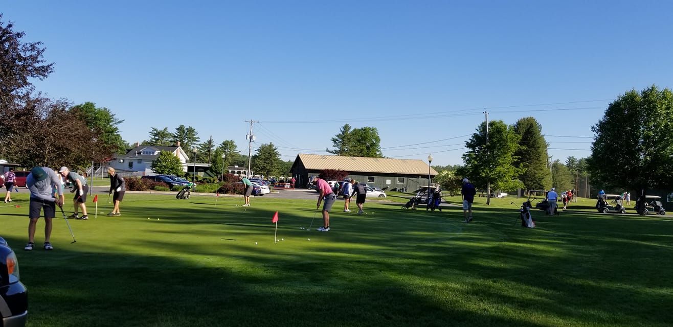 A group of people practicing putting on a sunlit golf course putting green with a building in the background.