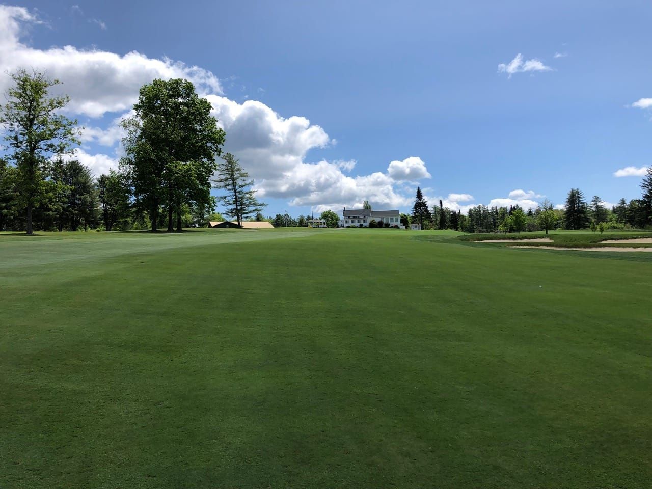 A sunny view of a green golf course fairway with a white building and trees in the background under a blue, cloudy sky.