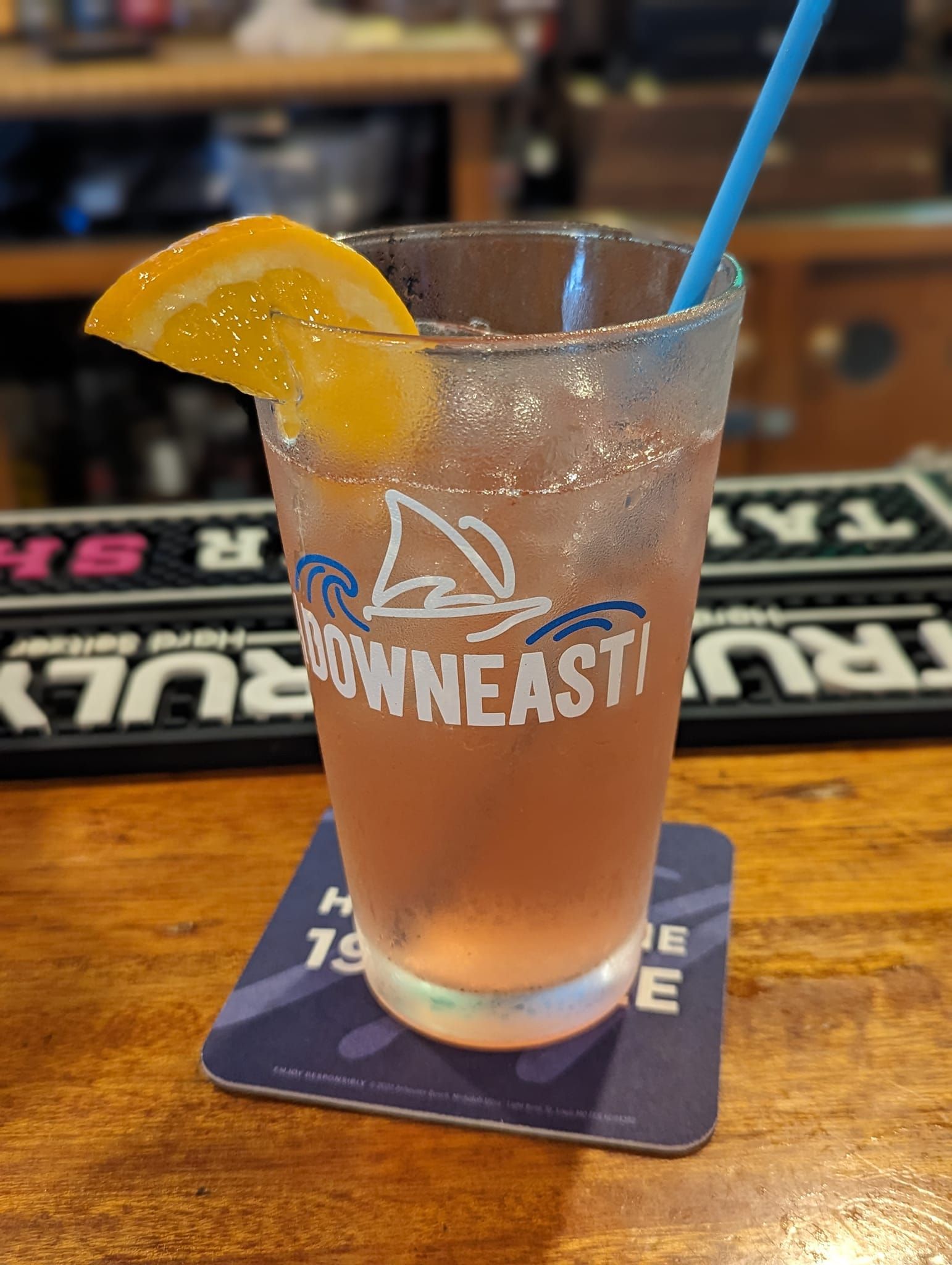 Cocktail in a glass with orange slice, blue straw, sitting on a coaster on a bar.