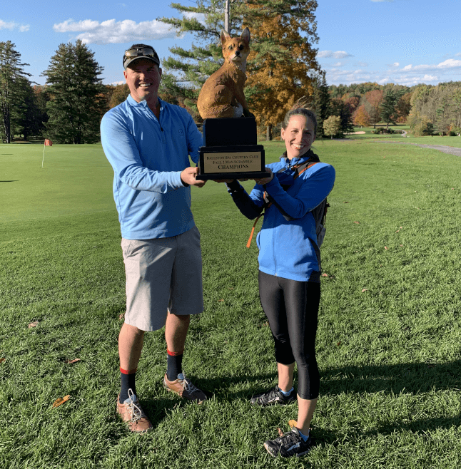 Man and woman holding a golf trophy with a fox statue on a green golf course.