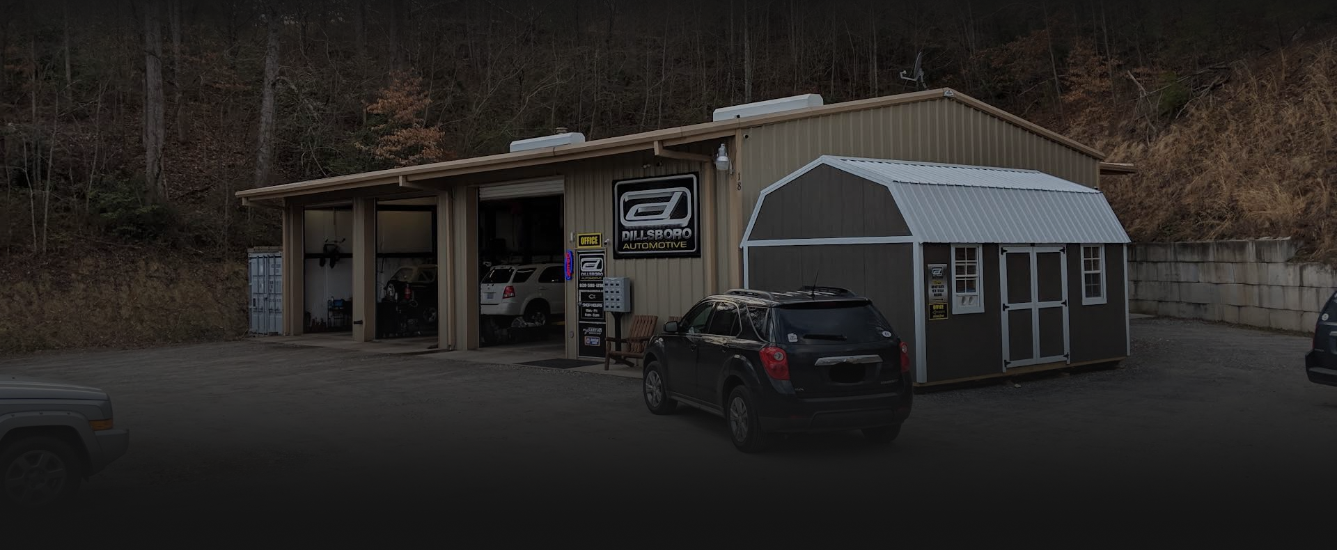 Gray shed-style building with an awning and parked SUV in a wooded lot | Dillsboro Automotive
