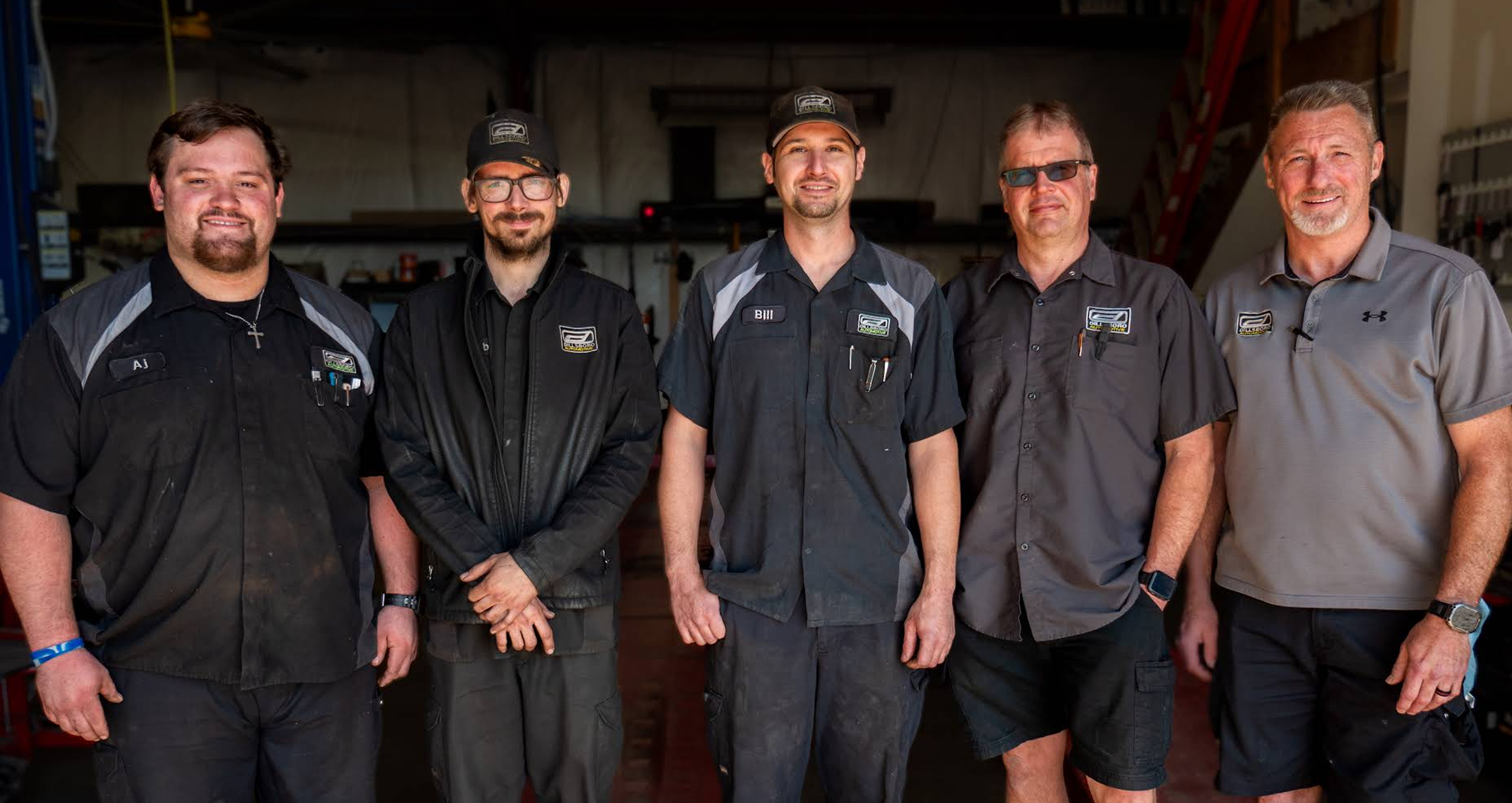 Five auto technicians standing in a garage, wearing black and gray shop uniforms. | Dillsboro Automotive