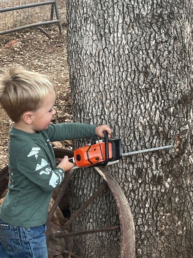 Young child pretending to cut a tree with an orange chainsaw, smiling. Next to an old wagon wheel.