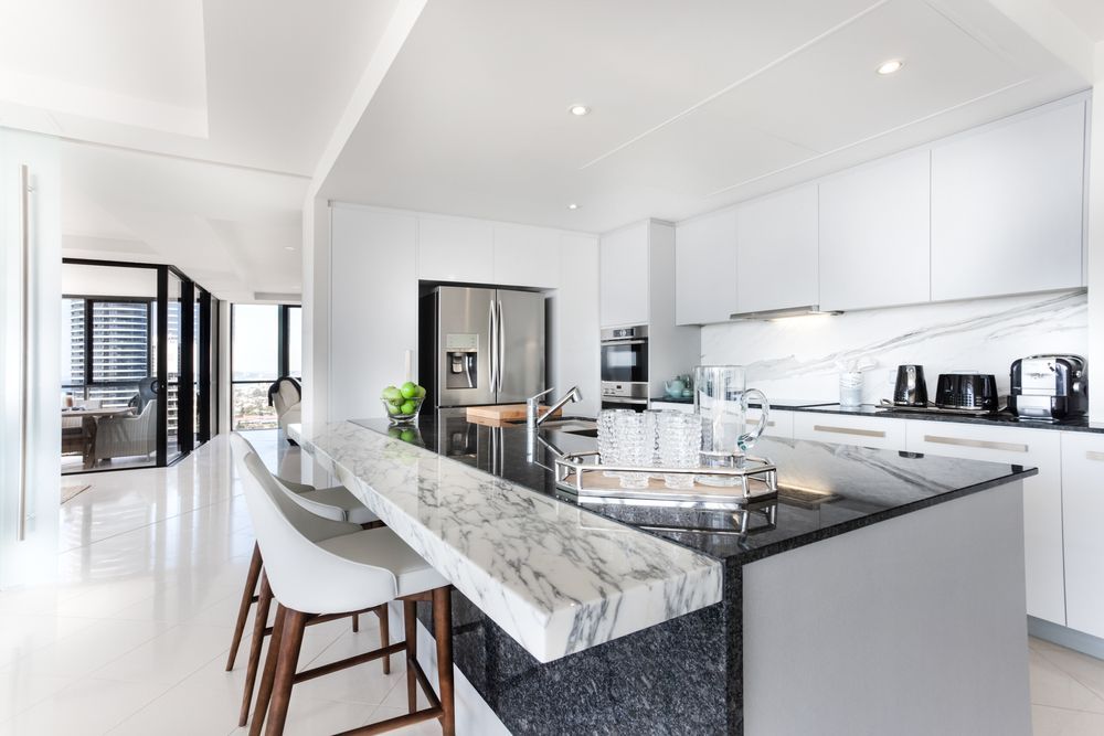 Modern, all-white kitchen with marble island and bar stools, stainless steel appliances, and open doorway — Verastone In Fyshwick, ACT